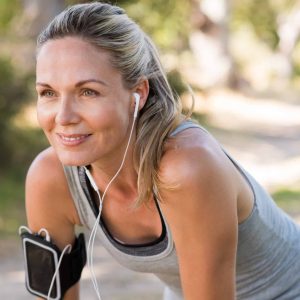 Portrait of athletic mature woman resting after jogging. Beautiful senior blonde woman running at the park on a sunny day. Female runner listening to music while jogging.