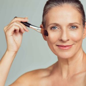 Closeup portrait of mature woman applying tonal foundation on face with makeup brush. Smiling senior woman applying powder for make up. Cosmetics and make-up portrait.